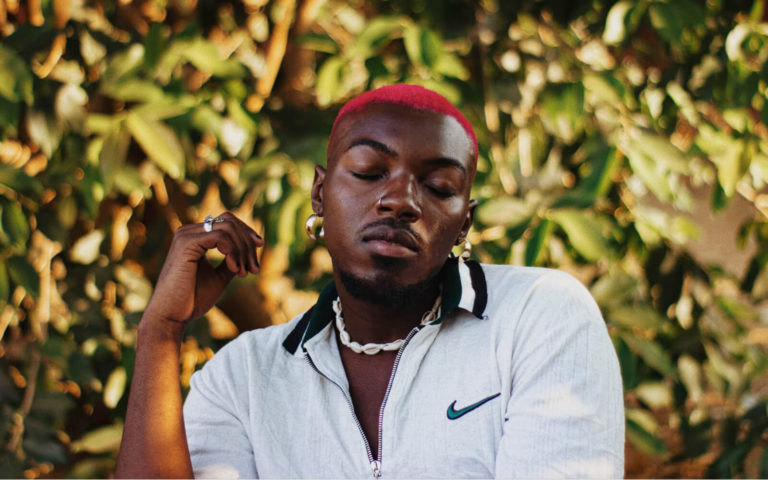 Zambian artist Bacci is sitting in a chair, his hair dyed the colour red and his eyes are closed.